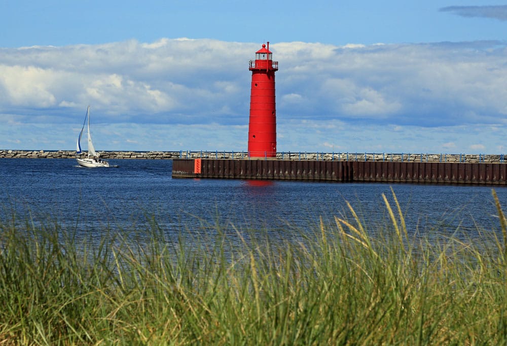Pere Marquette Lighthouse in Muskegon, Michigan, with a sailboat in the background and beach grass in the foreground.